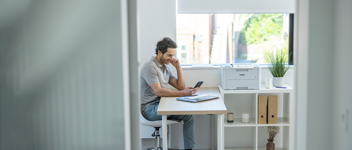 Man sat on chair at desk looking at his phone next to a window with a printer, folders and plants on shelving