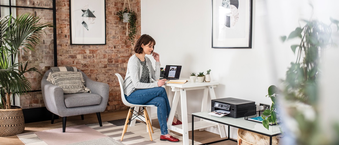 Woman sat at desk at home looking at her notepad pad and laptop with printer, plants and a chair in the foreground