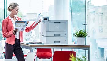 Woman stood picking document from printer, table, plant, red chairs, large windows