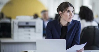 Woman sat at desk holding paper, laptop, printer, office