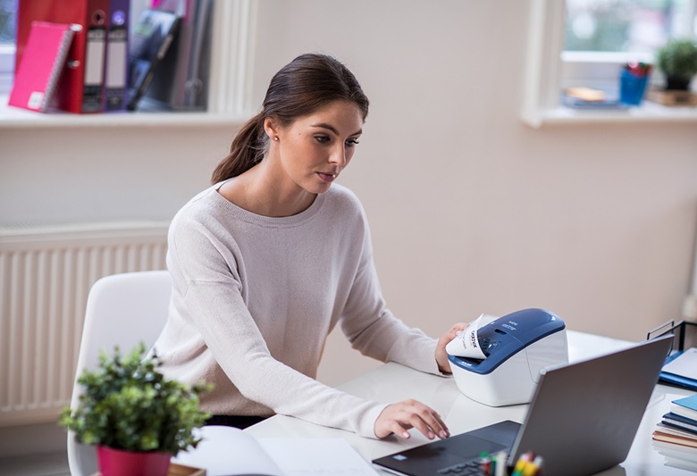 Woman sat looking at laptop printing a label from a device on her left.